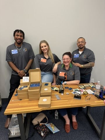 Four people smiling behind a table with small boxes and products on display at an event, all wearing matching gray shirts with logos.
