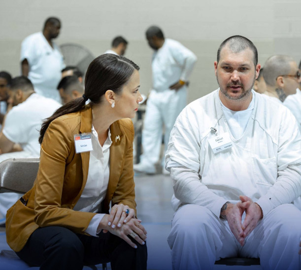A woman in a brown blazer sits and talks with a man in a white uniform during a Prison Entrepreneurship Program session, with others in white uniforms gathered in the background.