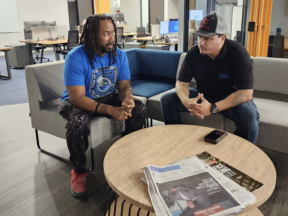 Two PEP participants engaged in a focused one-on-one conversation in a modern workspace, sitting across from each other with newspapers and a phone on the table.