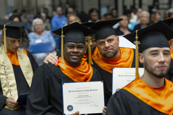 Graduates in caps and gowns smile and hold up their Prison Entrepreneurship Program certificates during a ceremony.