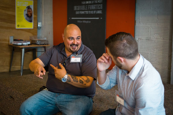 Two men sit indoors having a lively conversation, with one man pointing animatedly while the other listens and smiles.