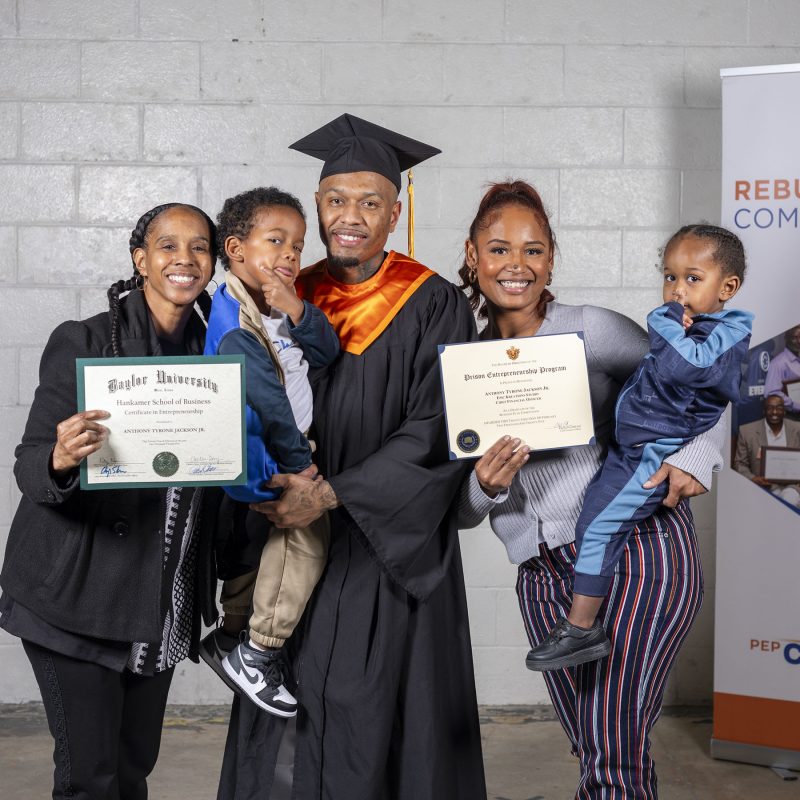 PEP graduate in cap and gown holding a young child, standing proudly with family members holding certificates and celebrating his achievement.