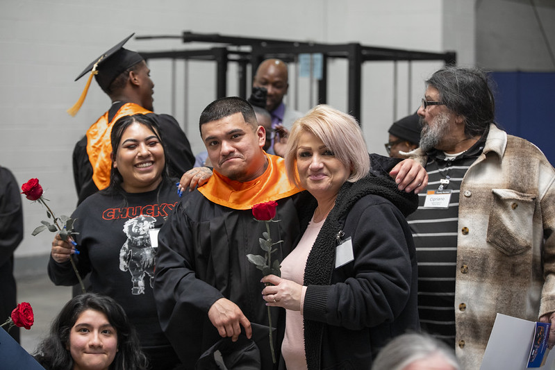 PEP graduate in cap and gown celebrating with family members holding roses at graduation ceremony.