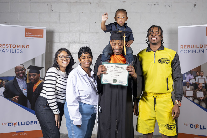 PEP graduate in cap and gown holding diploma, celebrating with family including child on shoulders, standing in front of PEP banners.