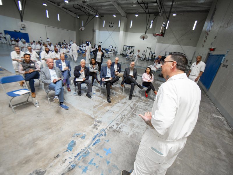 PEP participant in white uniform presents a business pitch to a panel of volunteers seated in a prison gymnasium while other incarcerated men observe from the background.