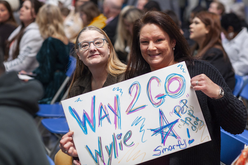 Supporters in the audience holding a colorful handmade sign that reads "Way 2 Go Wylie" at a PEP graduation event.