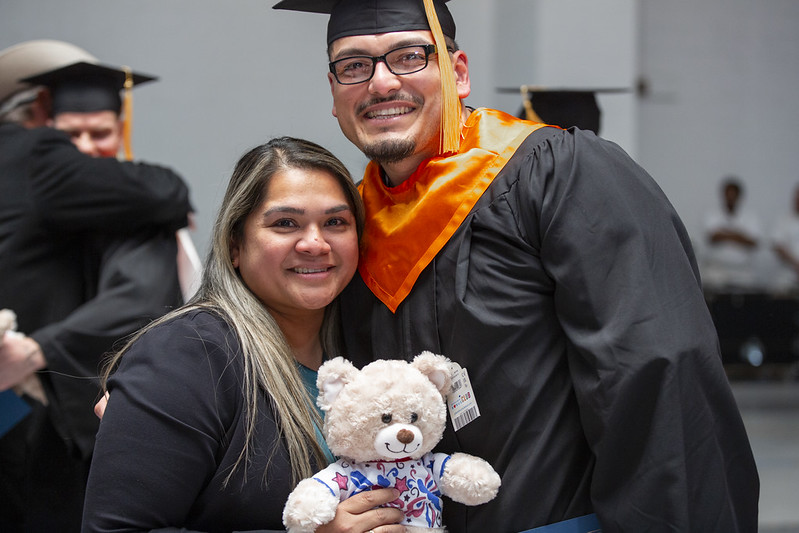 ChatGPT said: Smiling graduate in cap and gown with orange stole posing with a woman holding a teddy bear during a Prison Entrepreneurship Program graduation celebration.