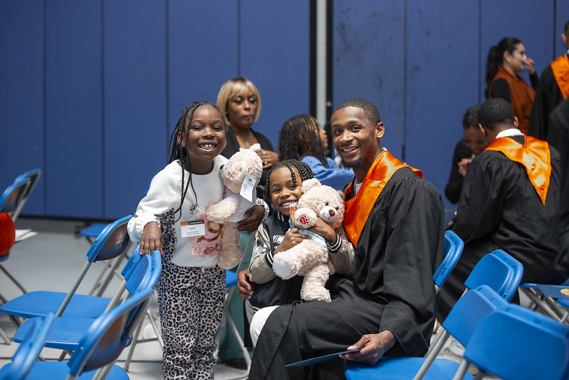 PEP graduate in cap and gown smiling with two young children holding teddy bears during the graduation celebration.