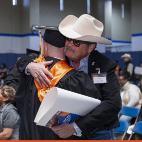 PEP graduate in cap and gown with orange stole hugging a supporter in a cowboy hat and sunglasses during graduation celebration.