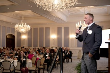 Man in a blazer speaking into a microphone on stage at a PEP event, with an audience seated at round tables in a ballroom with chandeliers.