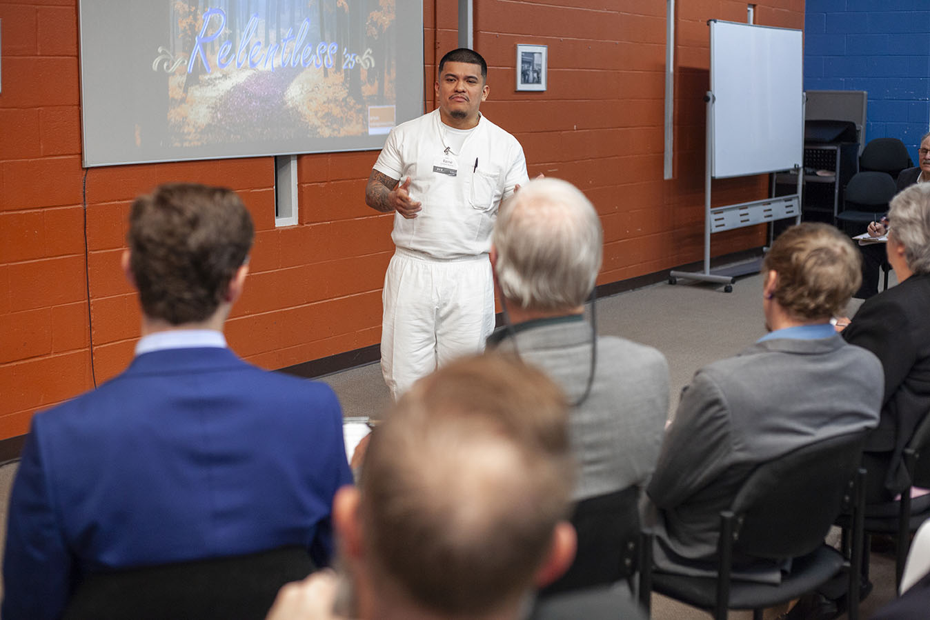 A man in a white uniform is giving a presentation to an audience seated in front of him. A slide projected on the screen behind him displays the word "Relentless."